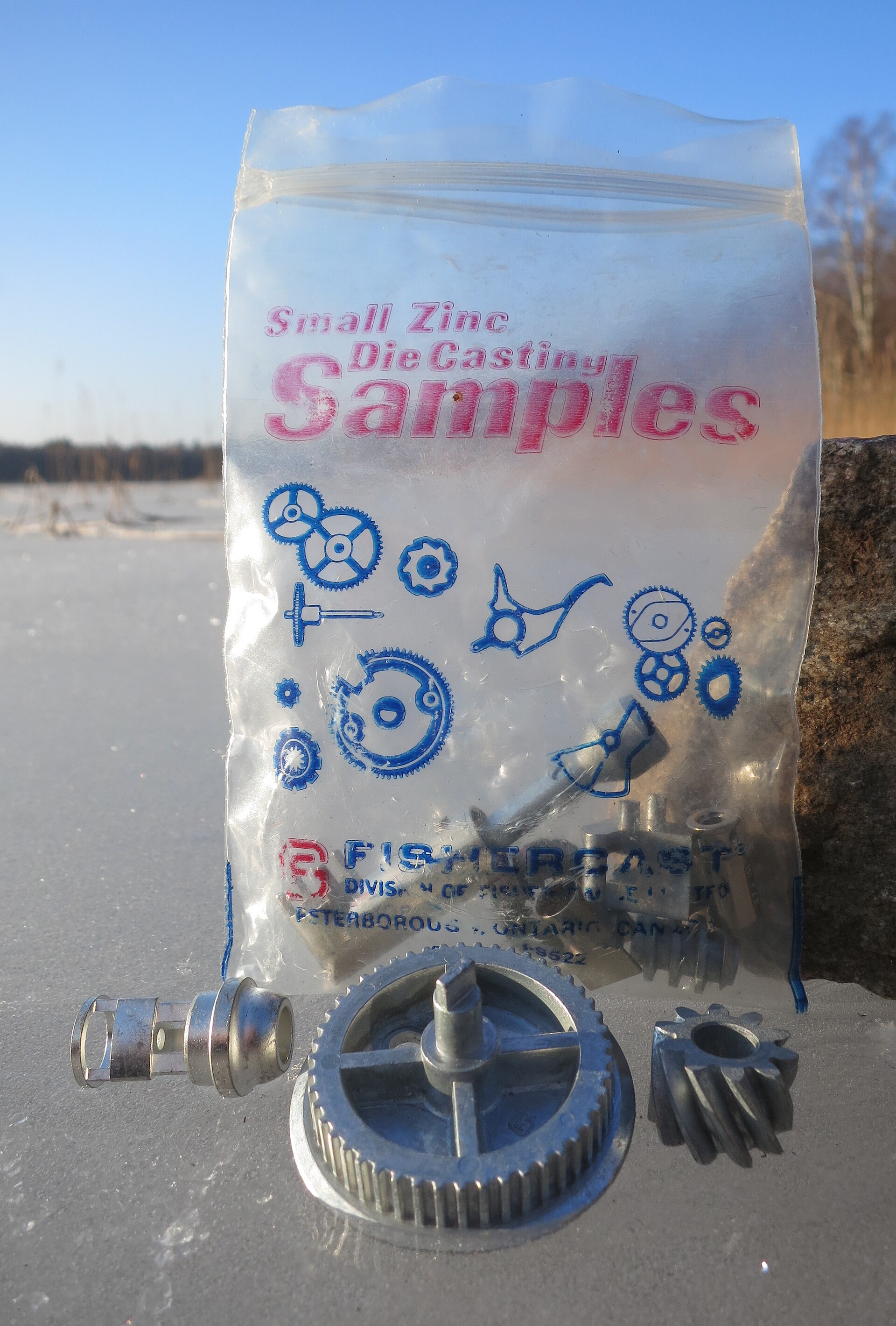 Small zinc die-cast sample parts on ice beside a Fishercast sample bag